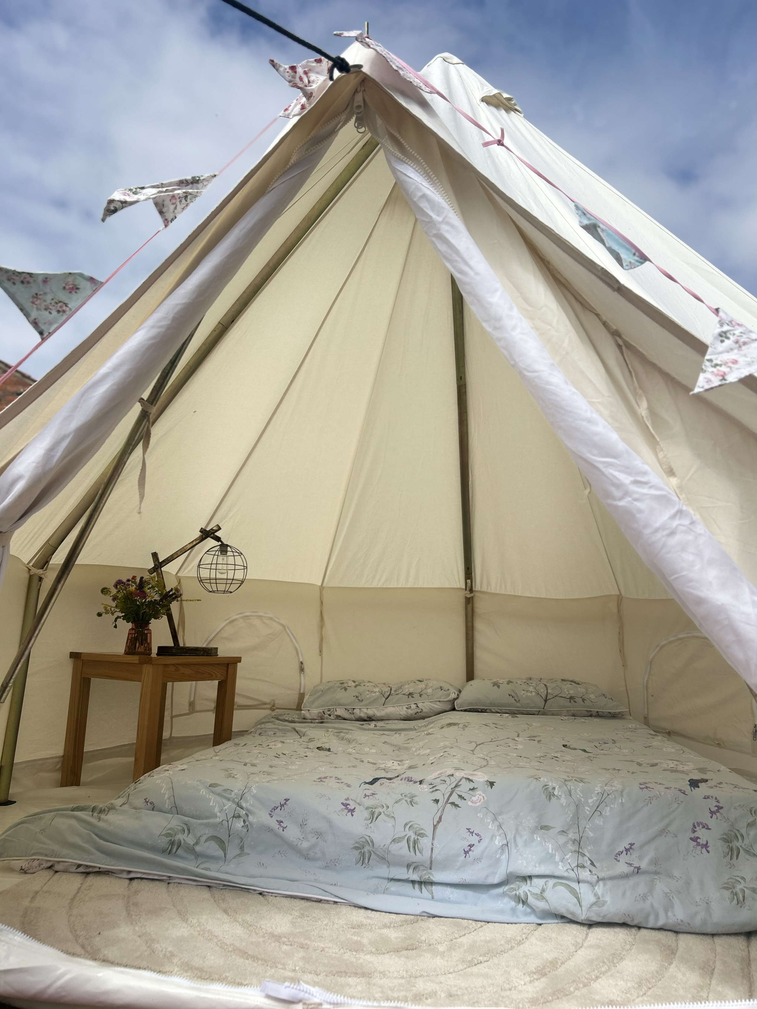 View of the interior of a cream-colored canvas tent with a bed that has floral-patterned bedding, a small wooden side table with a flower vase, and bunting hanging along the ceiling. The tent is set up outside under a partly cloudy sky.