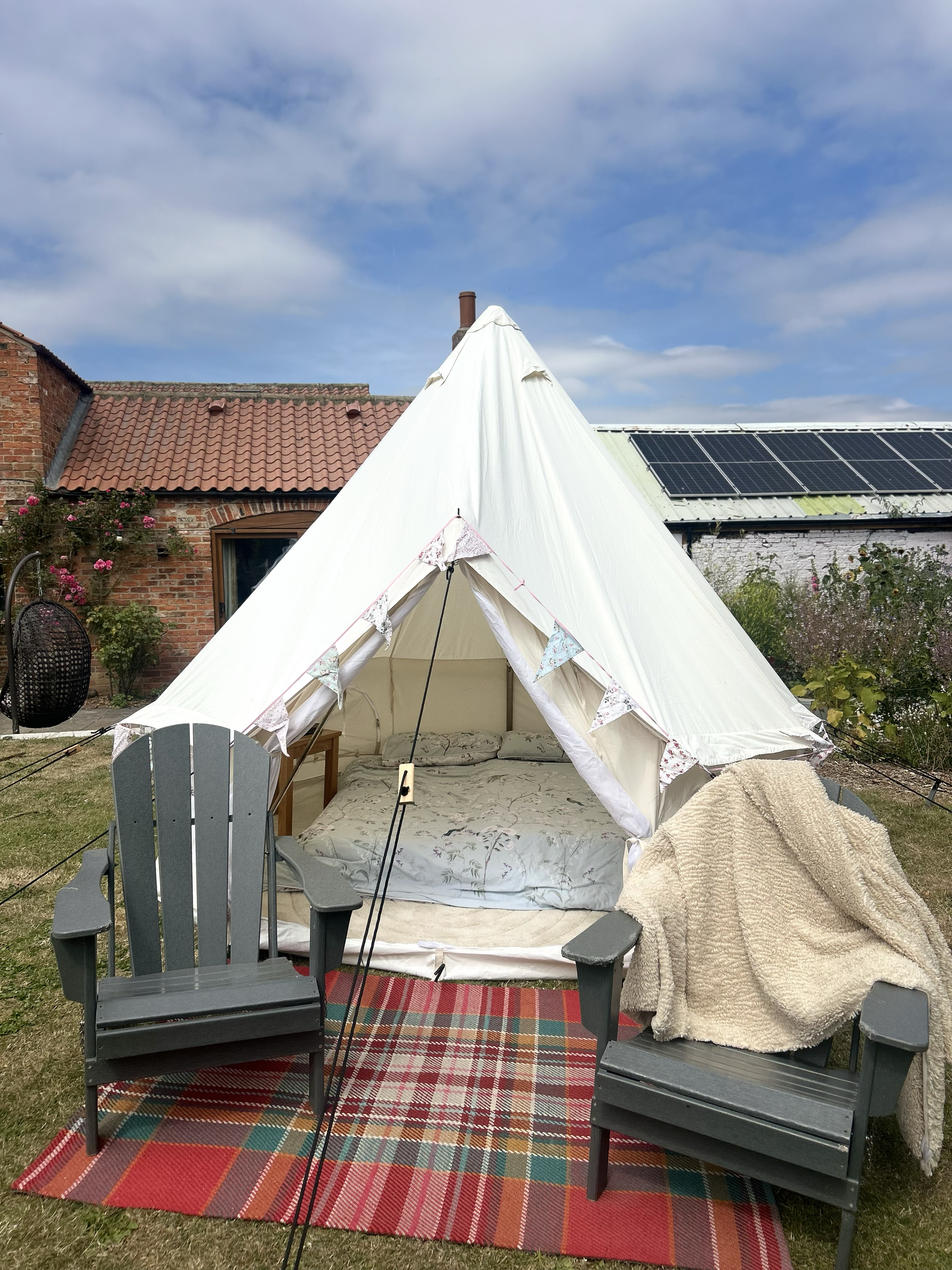 A backyard scene with a white canvas teepee tent, two gray Adirondack chairs, a colorful plaid outdoor rug, and various garden plants and flowers. There are two black lanterns, a small wooden table, and a towel draped over one of the chairs. The back