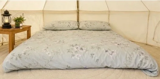 A bed with floral bedding and two pillows inside a tent or canopy with a beige fabric wall, a wooden nightstand, a lantern, and a small plant.