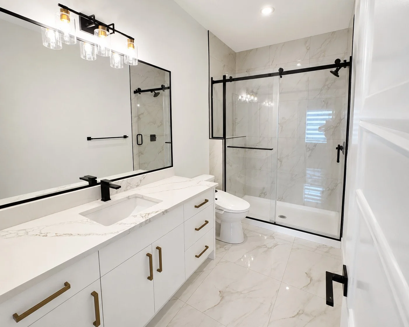 Modern bathroom with white marble floor and walls, black fixtures, a large wall mirror, a white vanity with a marble countertop and gold handles, a glass-enclosed shower with black framing, and a white toilet.