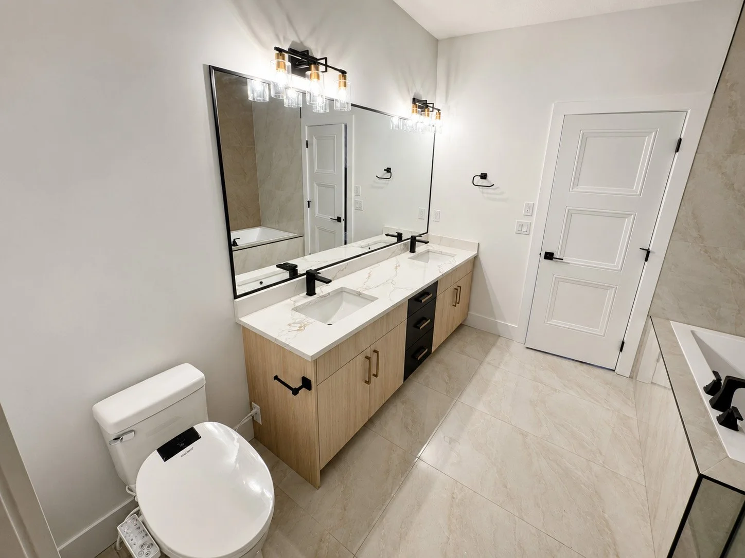 Modern bathroom with double vanity featuring white marble countertop, two black faucets, large mirror, light fixtures, and beige tiled floor.