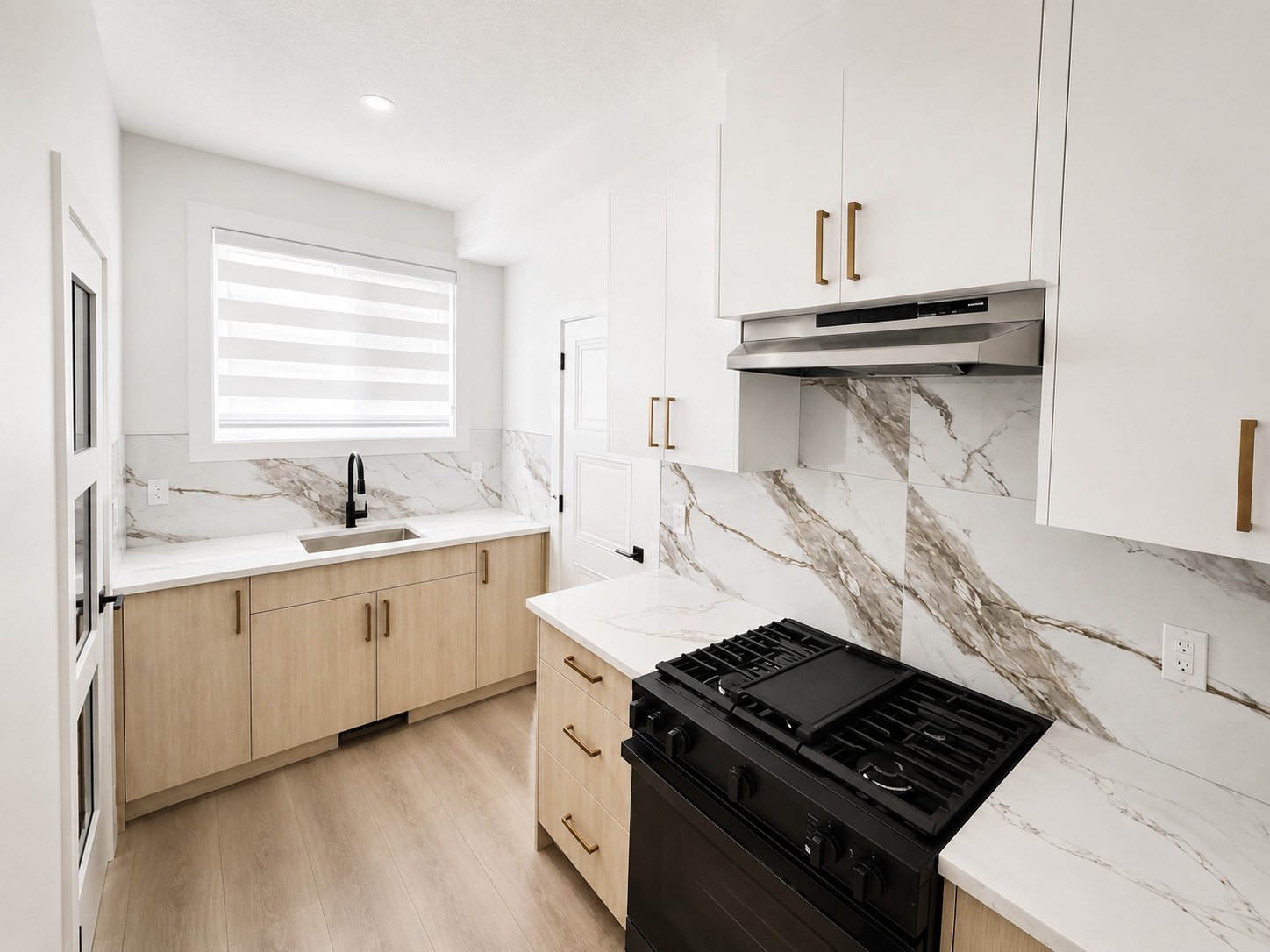 Modern kitchen with white upper cabinets, light wood lower cabinets, marble backsplash, black stove, and a window with a striped roller shade.