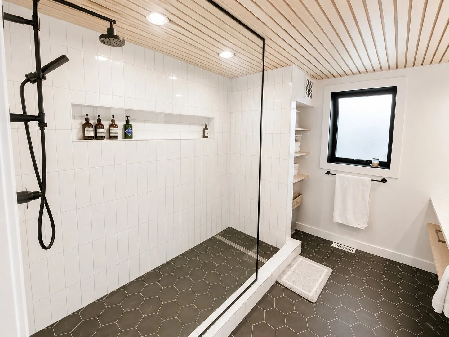 Modern bathroom with walk-in shower enclosed by a glass partition, white tiles, black hexagonal floor tiles, wooden ceiling, and a frosted window.