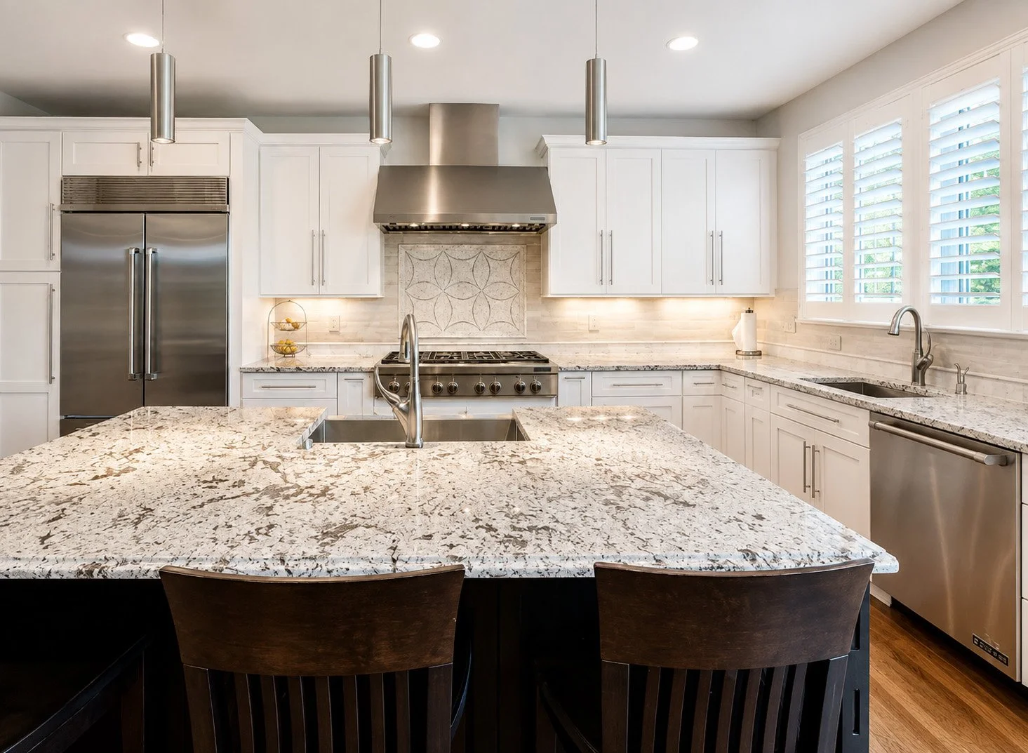 Modern kitchen with white cabinets, granite countertops, a central island with a double sink, stainless steel appliances, and large windows with white shutters.