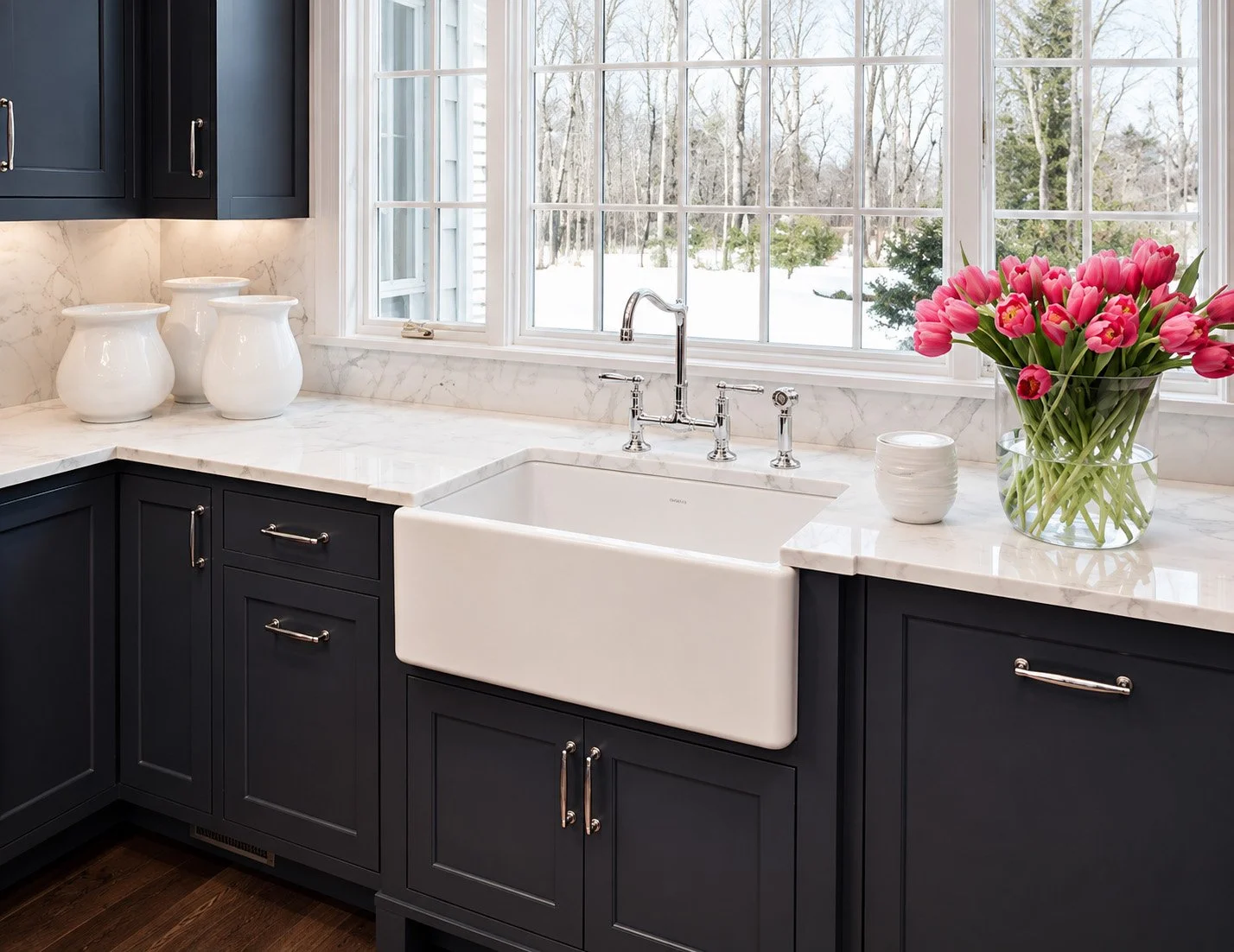 Kitchen with white farmhouse sink, navy blue cabinets, white marble countertops, large window with a snowy landscape outside, pink tulips in a glass vase, white ceramic jars, and small white jar on the counter.