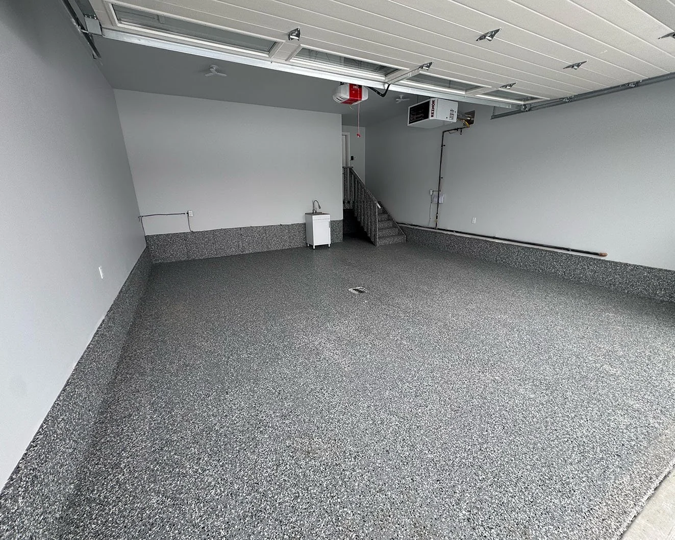 Empty garage with speckled gray floors, white walls, a small sink, a staircase, and garage door openers.