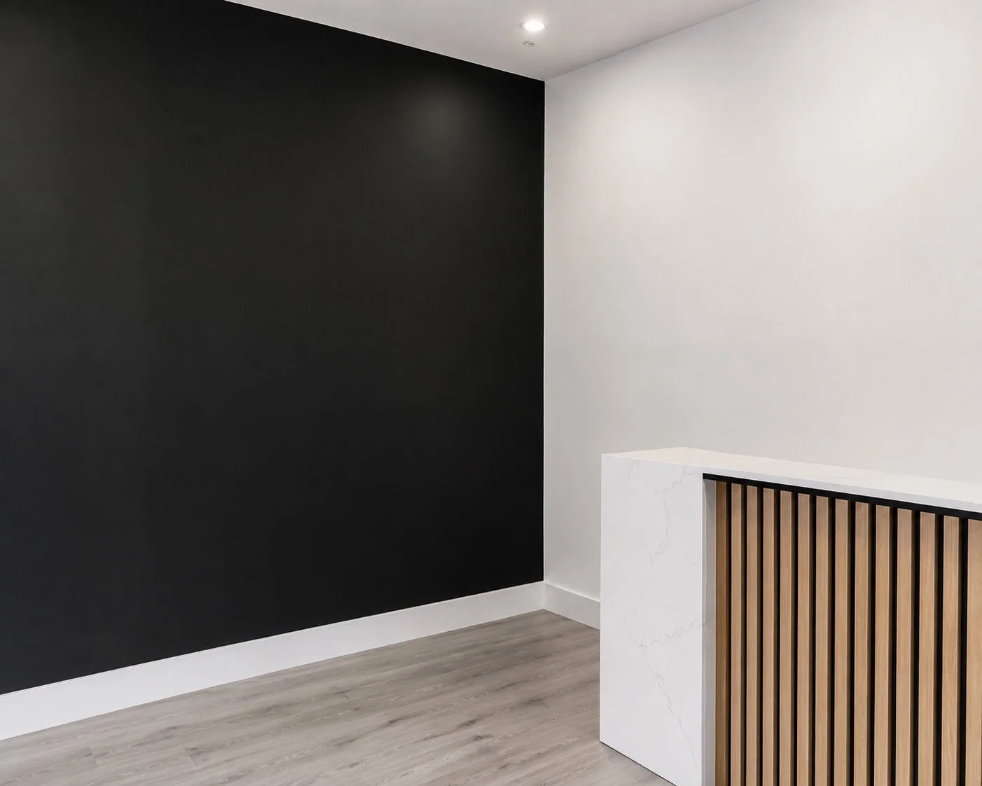 A modern interior corner with black and white walls, a wooden slat design on a white marble counter or partition, and a wood laminate floor.