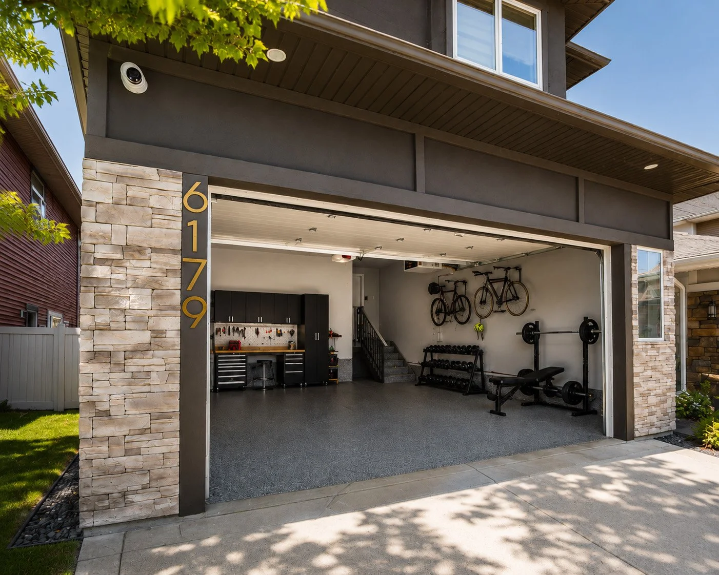 The garage of a modern house with the door open, showing exercise equipment including a barbell and weights, bicycles hanging on the wall, black storage cabinets, and a pegboard with tools on the wall.