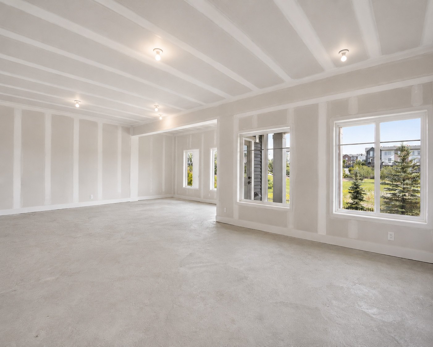 Empty and unfinished white room with large windows showing outdoor trees and neighborhood, ceiling with exposed beams and multiple light fixtures.