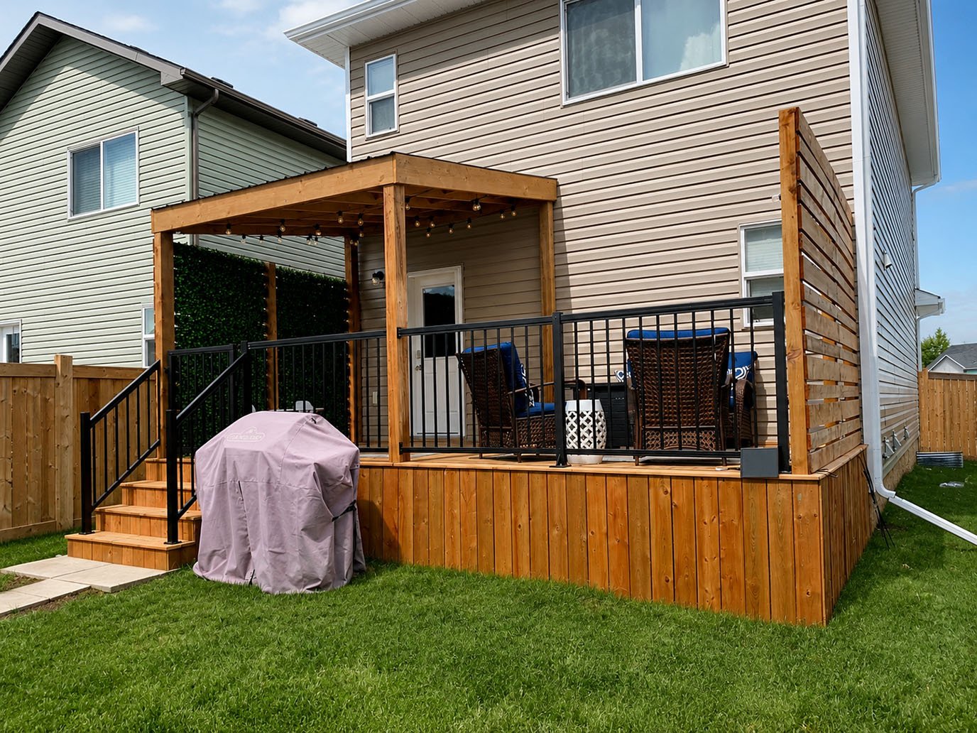 A backyard deck attached to a beige house, with stairs leading down to a grassy lawn. The deck has a black metal railing, outdoor furniture including chairs and a small table, and a covered grill. There is a wooden privacy wall and string lights hang