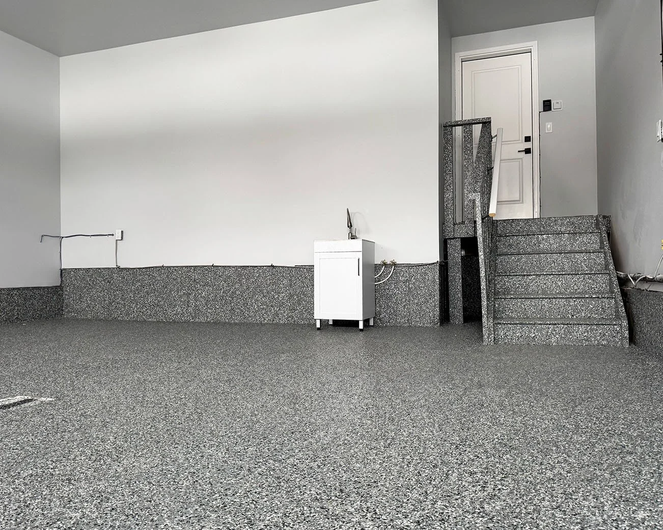 Empty garage with gray speckled flooring, a white wall with black electrical cables, a white utility sink, and gray stairs leading to a white door.