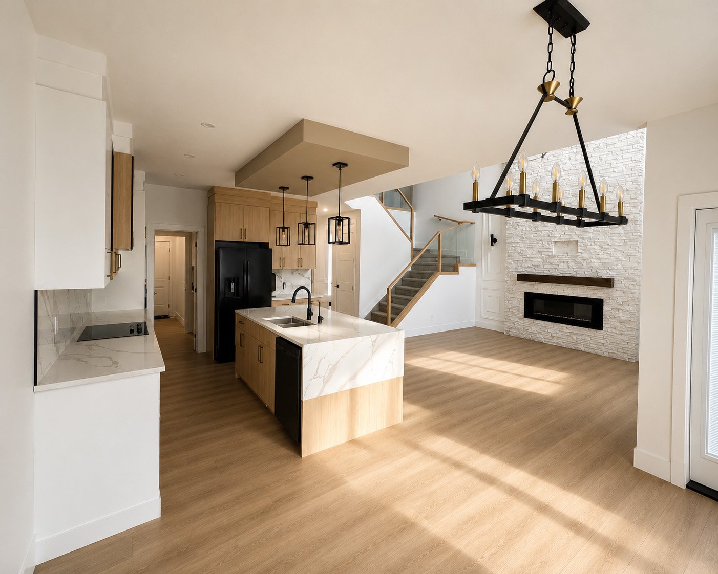 Open-concept living room and kitchen with wood flooring, white walls, modern fireplace with white textured stone surround, black fireplace insert, staircase with wooden handrail, and large black chandelier with candles.