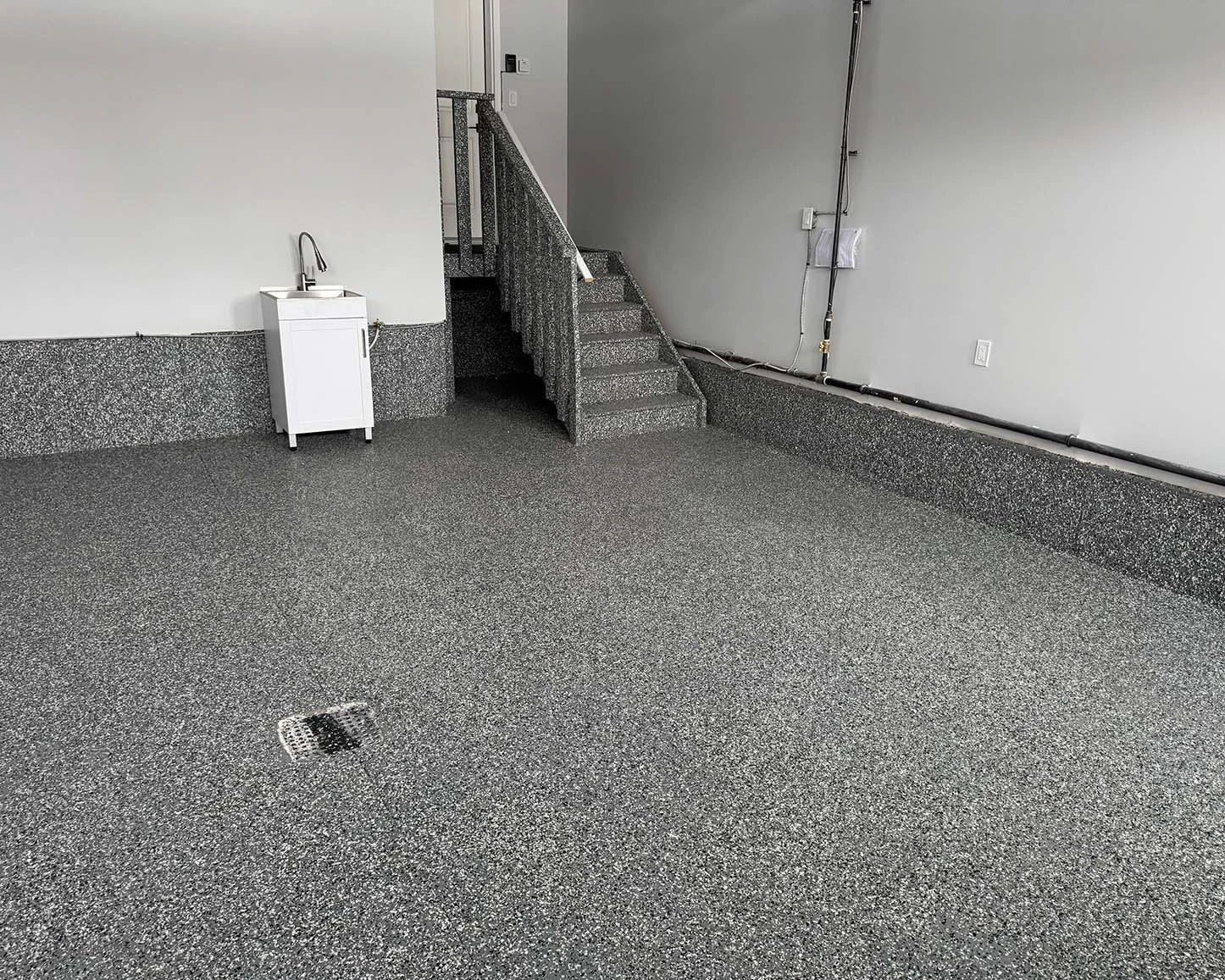 Empty room with speckled gray epoxy floors, a small white utility sink, a staircase with speckled gray steps, and exposed electrical wiring along the wall.