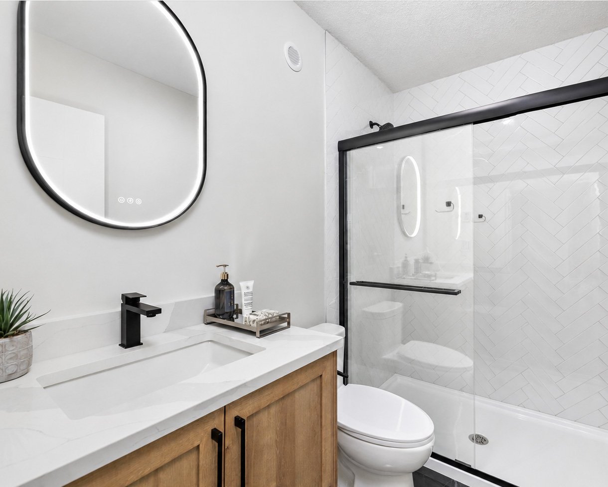 Modern bathroom with white vanity, black faucet, round mirror with LED light, potted plant, soap dispenser, bottle, and toiletries, toilet, and glass-enclosed shower with white herringbone tiles.