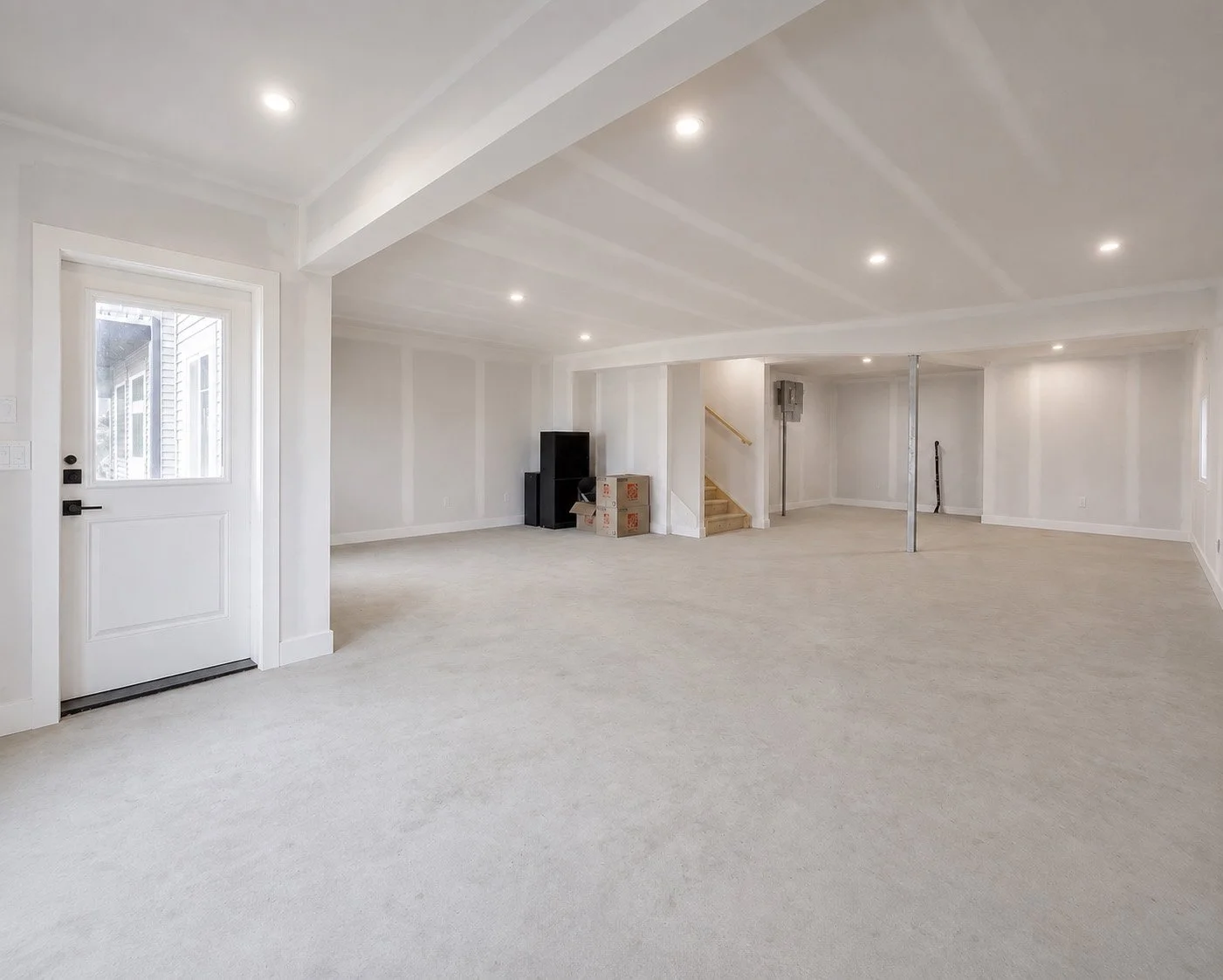 Empty finished basement with white walls, beige carpet, and recessed ceiling lights. A door to the outside is on the left, and a small staircase leads to an upper level. There are some boxes and black storage units near the back wall, with a metal pole and a pipe in the right back corner.