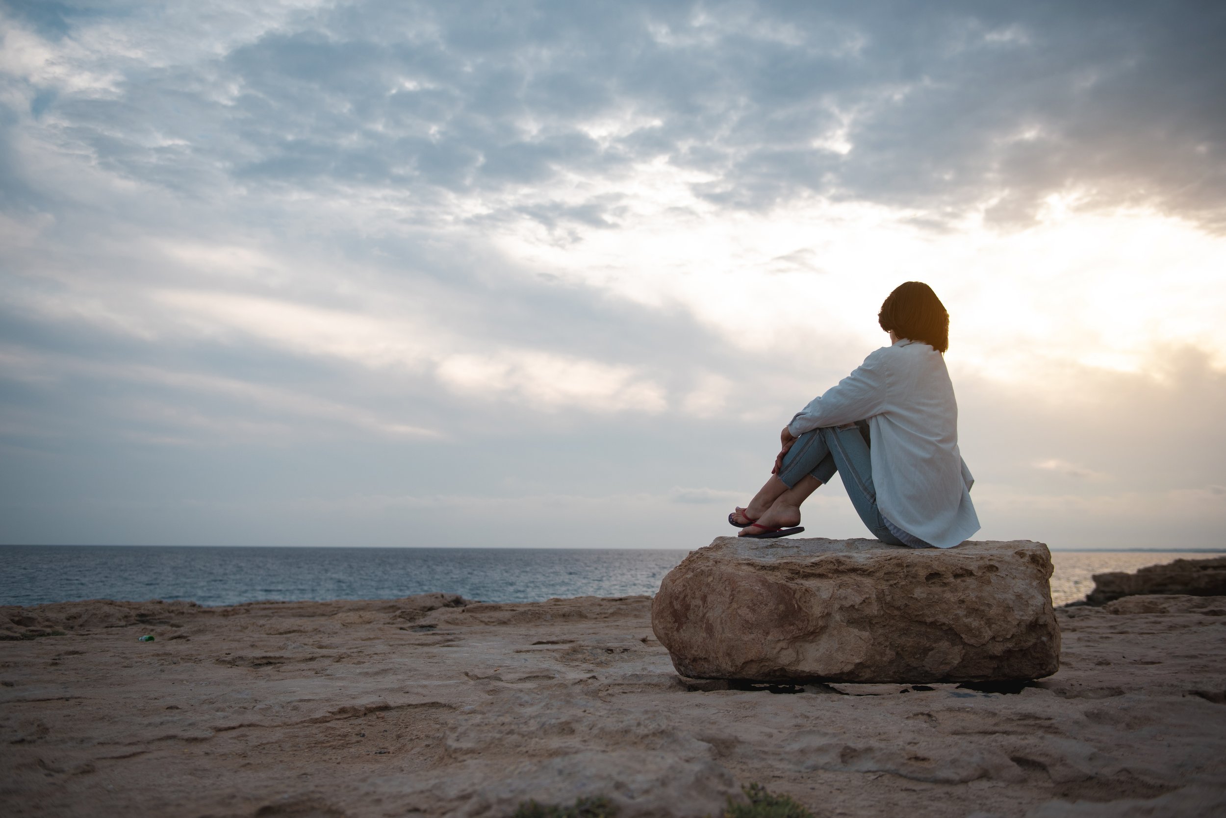 A woman sitting on a large rock by the ocean at sunset, looking at the sky with clouds.