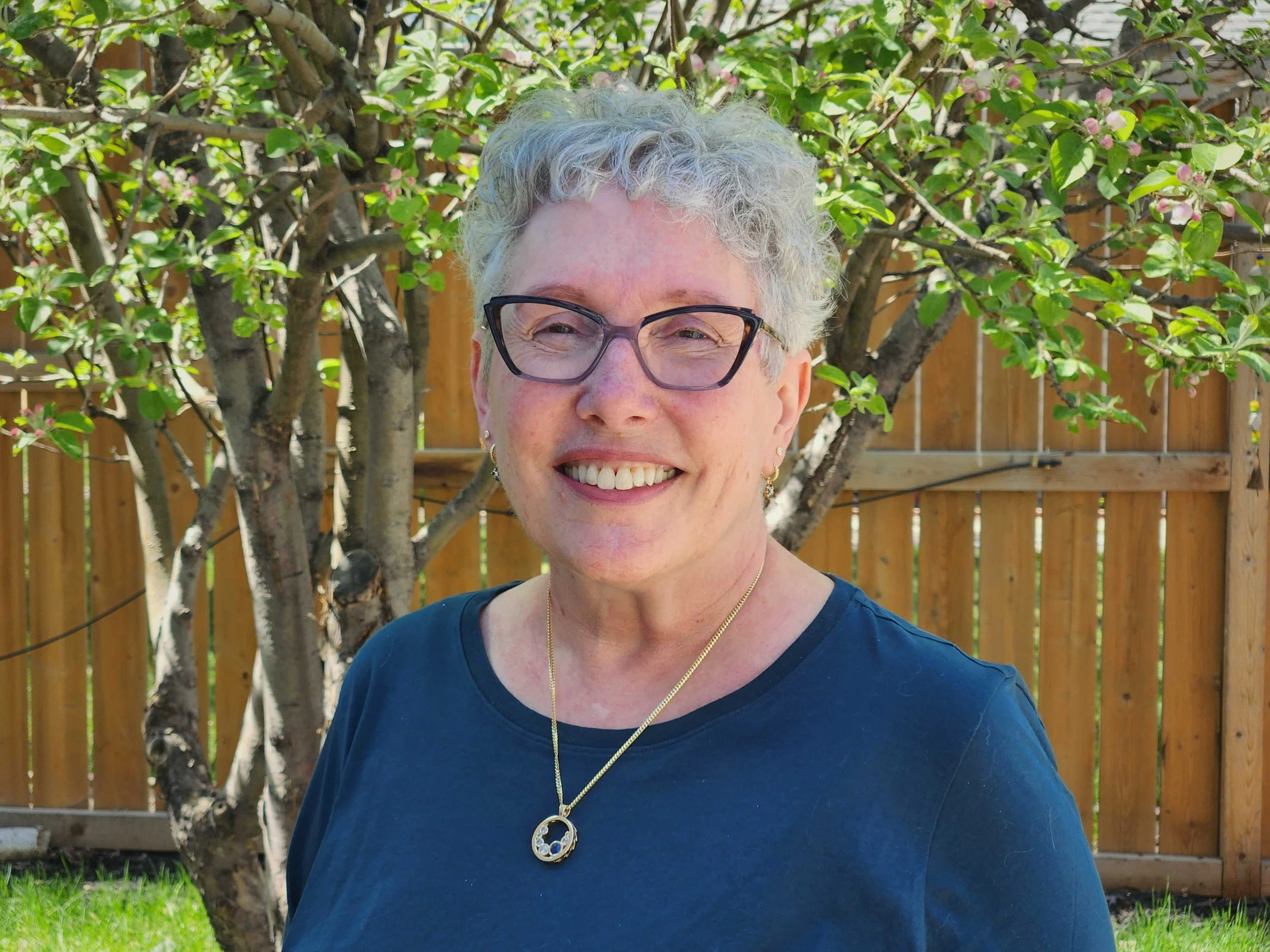 Kati Hatch smiling with short curly gray hair, wearing glasses, a navy blue top, and gold jewelry, standing outdoors in front of a tree with pink blossoms and a wooden fence.