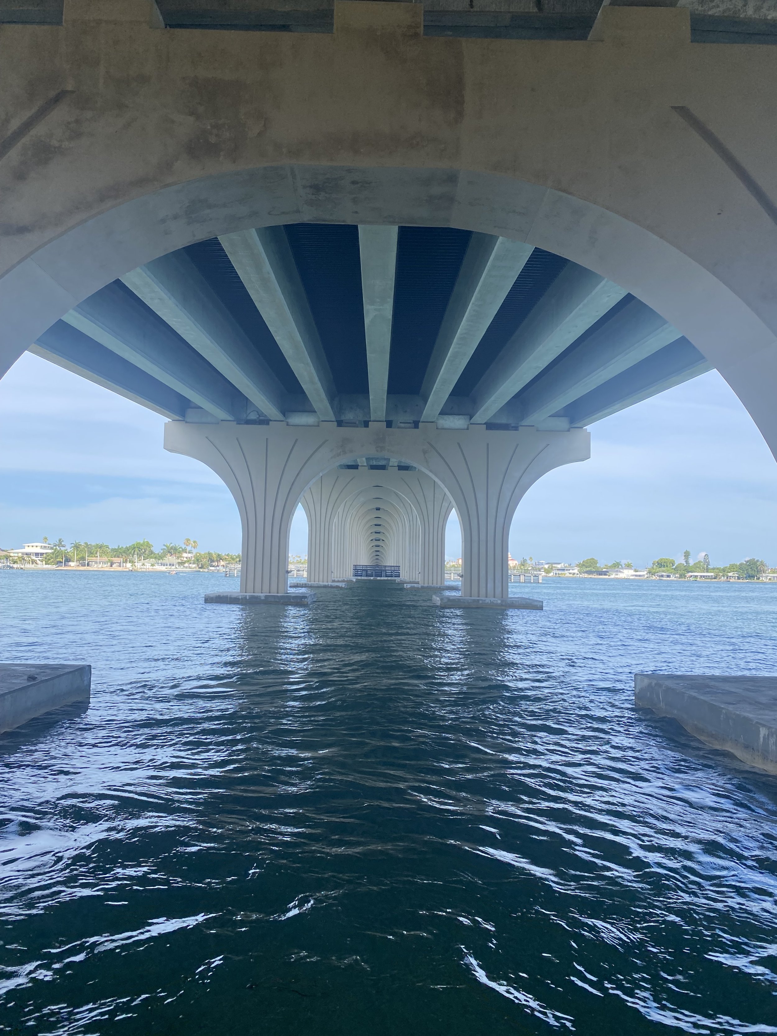 Underneath a large bridge with concrete arches and blue metal beams over a body of water with a cityscape in the background.