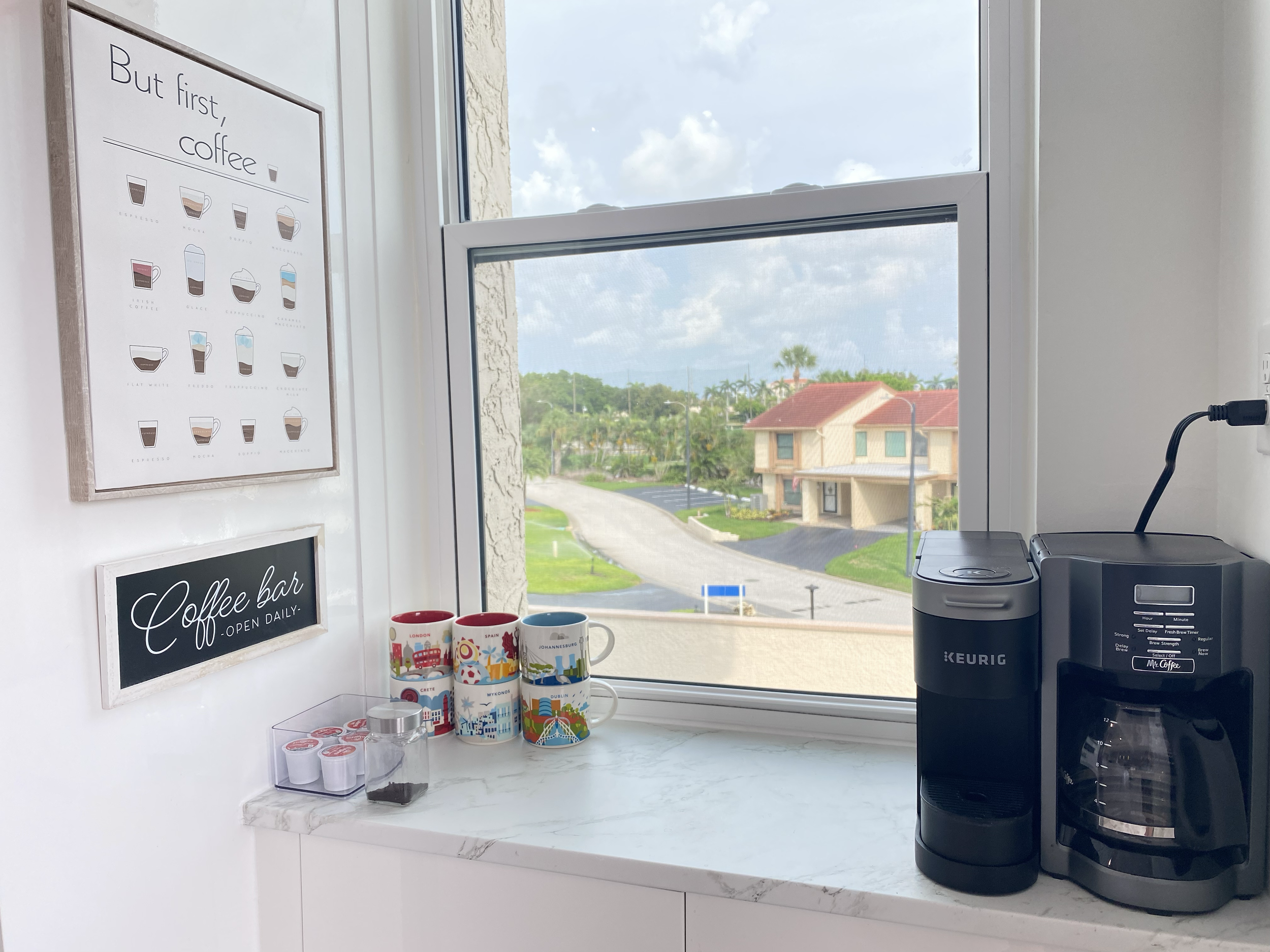 A coffee station with colorful souvenir mugs, a Keurig coffee maker, and a window showing a residential neighborhood with houses and palm trees.  For a luxury accomodation