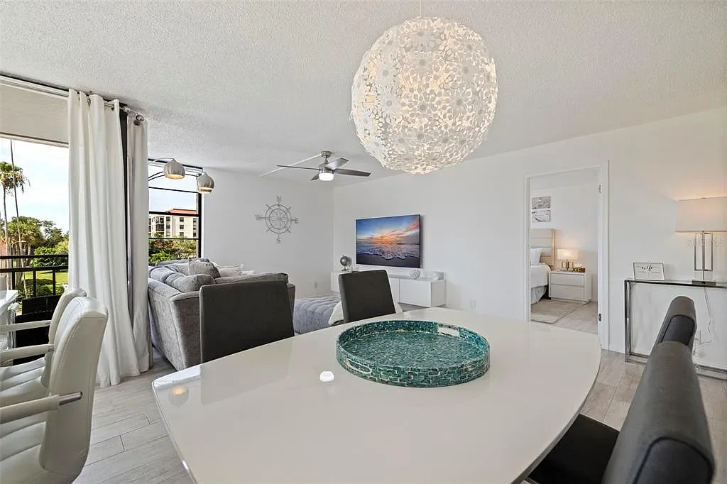 Contemporary living room and dining area with white walls, large window, gray and white furniture, a television, and a decorative pendant light over a white dining table with a green mosaic centerpiece For a luxury accomodation