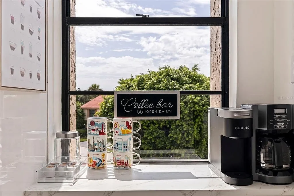 Coffee station with Keurig coffee maker, decorated mugs, glass jar with coffee pods, and a sign that reads 'Coffee Bar Open Daily' on a windowsill overlooking trees and a sky with clouds.  For a luxury accomodation