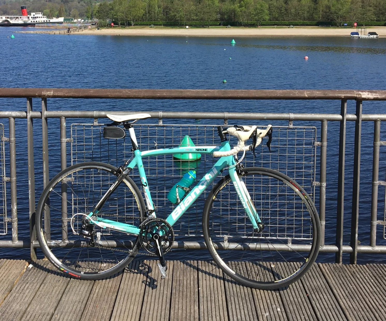 A light blue ale / carbon Bianchi road bicycle with water bottles, parked and locked to a metal railing on a wooden pier, overlooking a lake with buoys, boats, and a distant shoreline with trees. Complimentary of lightweight bike for our guests