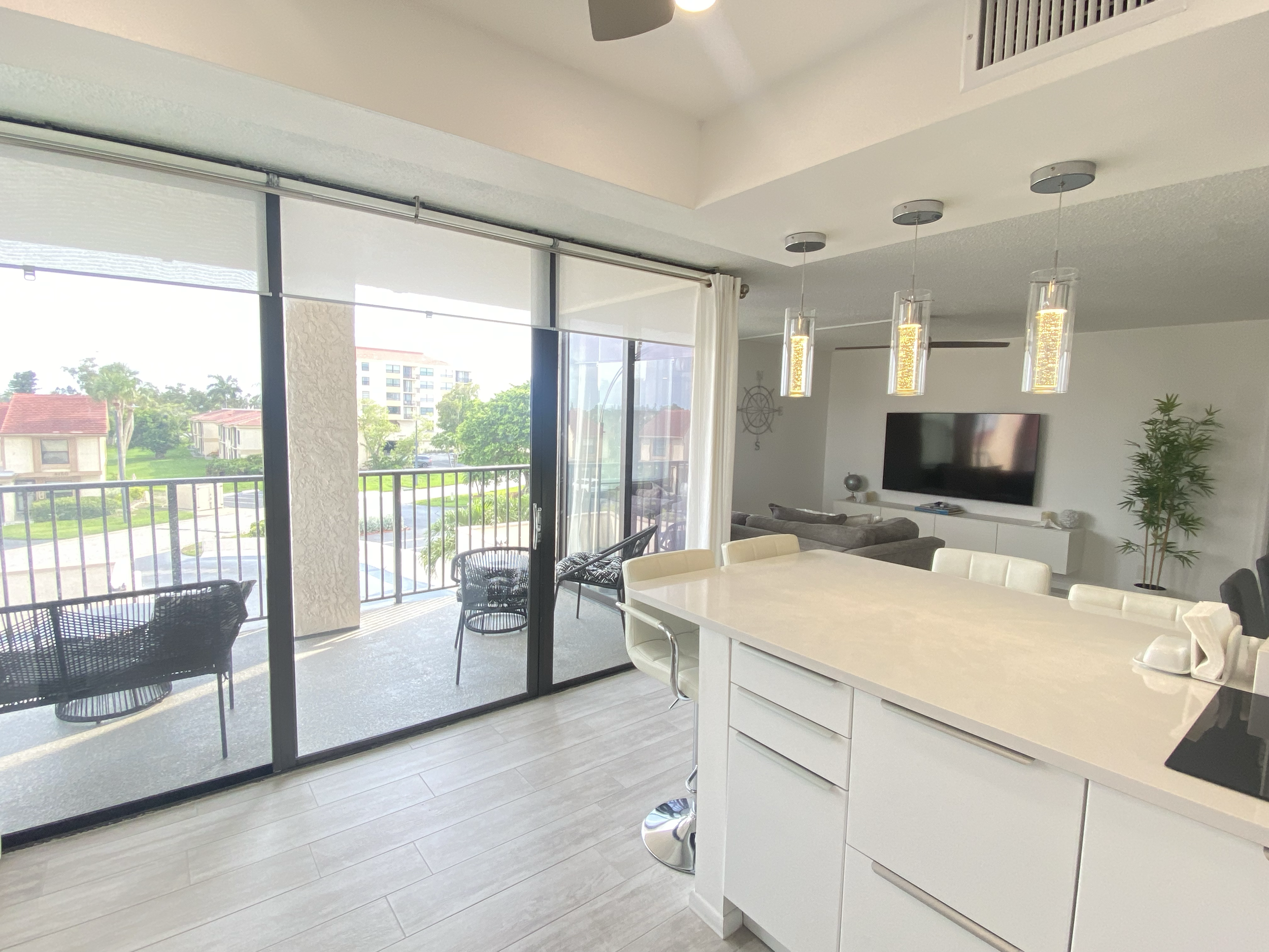 Open-concept living space with sliding glass doors leading to a balcony, view of neighboring buildings and greenery, modern decor including a TV wall, ceiling fan, dining table, and pendant lights. For a luxury experience