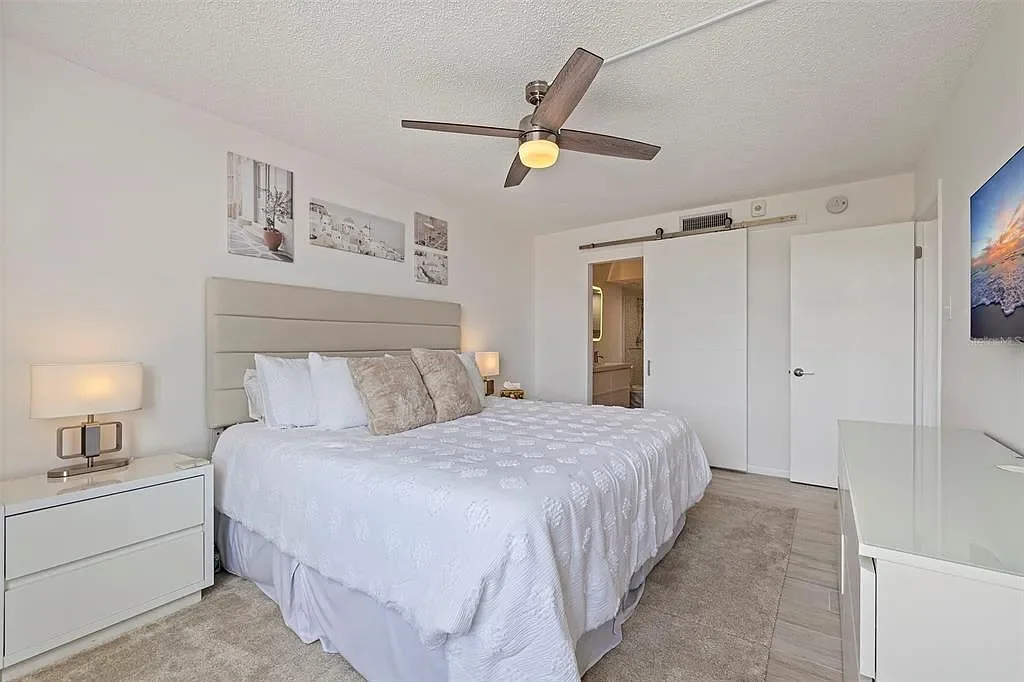 Modern bedroom with white bedding, beige headboard, two nightstands with lamps, ceiling fan, sliding closet doors, and a wall-mounted TV.For a luxury experience