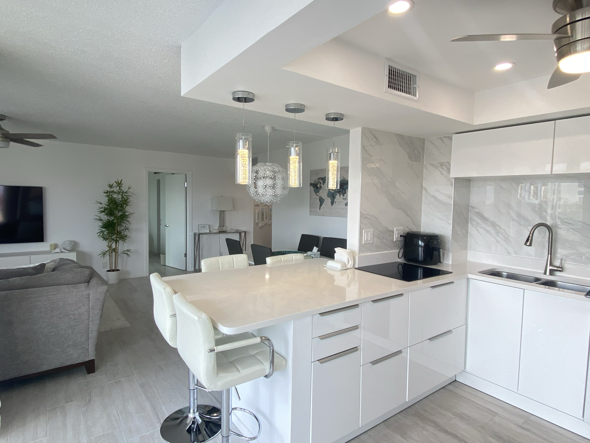 Modern open-concept kitchen with white cabinetry, marble backsplash, built-in stovetop, and a white island with barstools. The background shows a dining area with a table, chairs, a world map wall art, and pendant lighting.  For a luxury accomodation