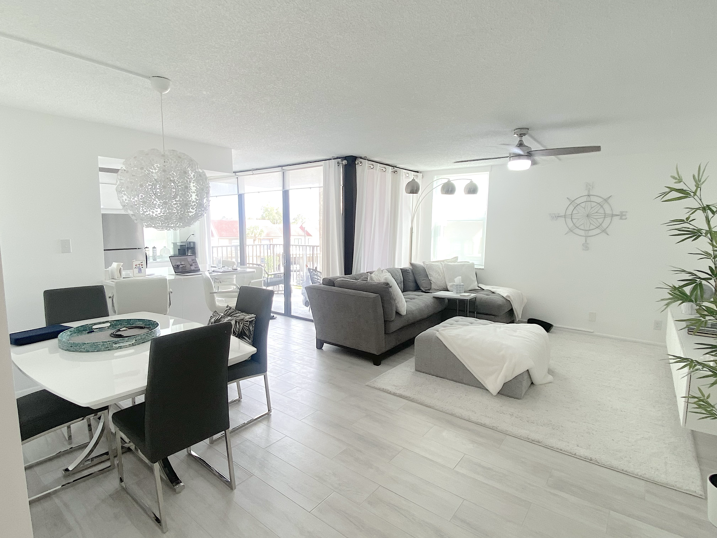 A spacious, modern and contemp living room with a dining area, a grey sectional sofa, a large sliding glass door leading to a balcony, white curtains, a hanging light fixture, a ceiling fan,  For a luxury experience