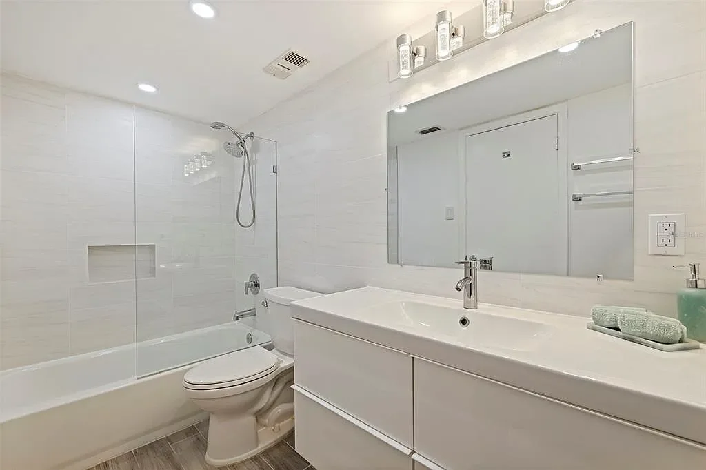 Modern bathroom with white tiles, bathtub with shower, toilet, large mirror, sink with faucet, and towel rack.  For a luxury accomodation