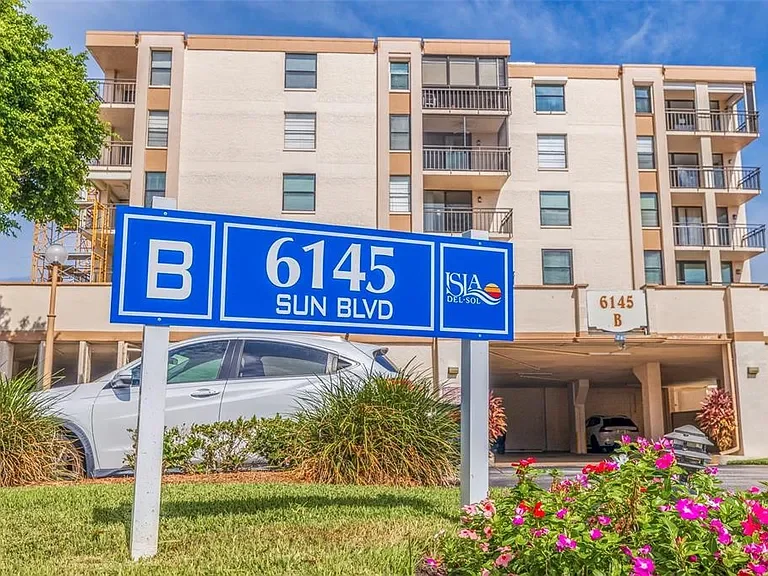 Apartment building with a parking garage, blue street sign in front reading 6145 Sun Blvd, with a car parked, flower bushes, and a clear blue sky. Beautiful gardens and flowers and well maintained grounds