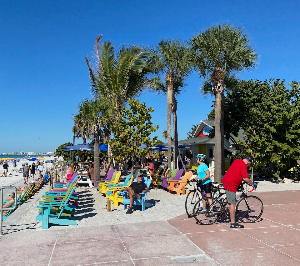 Beachside scene with colorful Adirondack chairs, people relaxing and socializing, palm trees, a small building, bicycles, and a sandy beach in the background under a clear blue sky.
