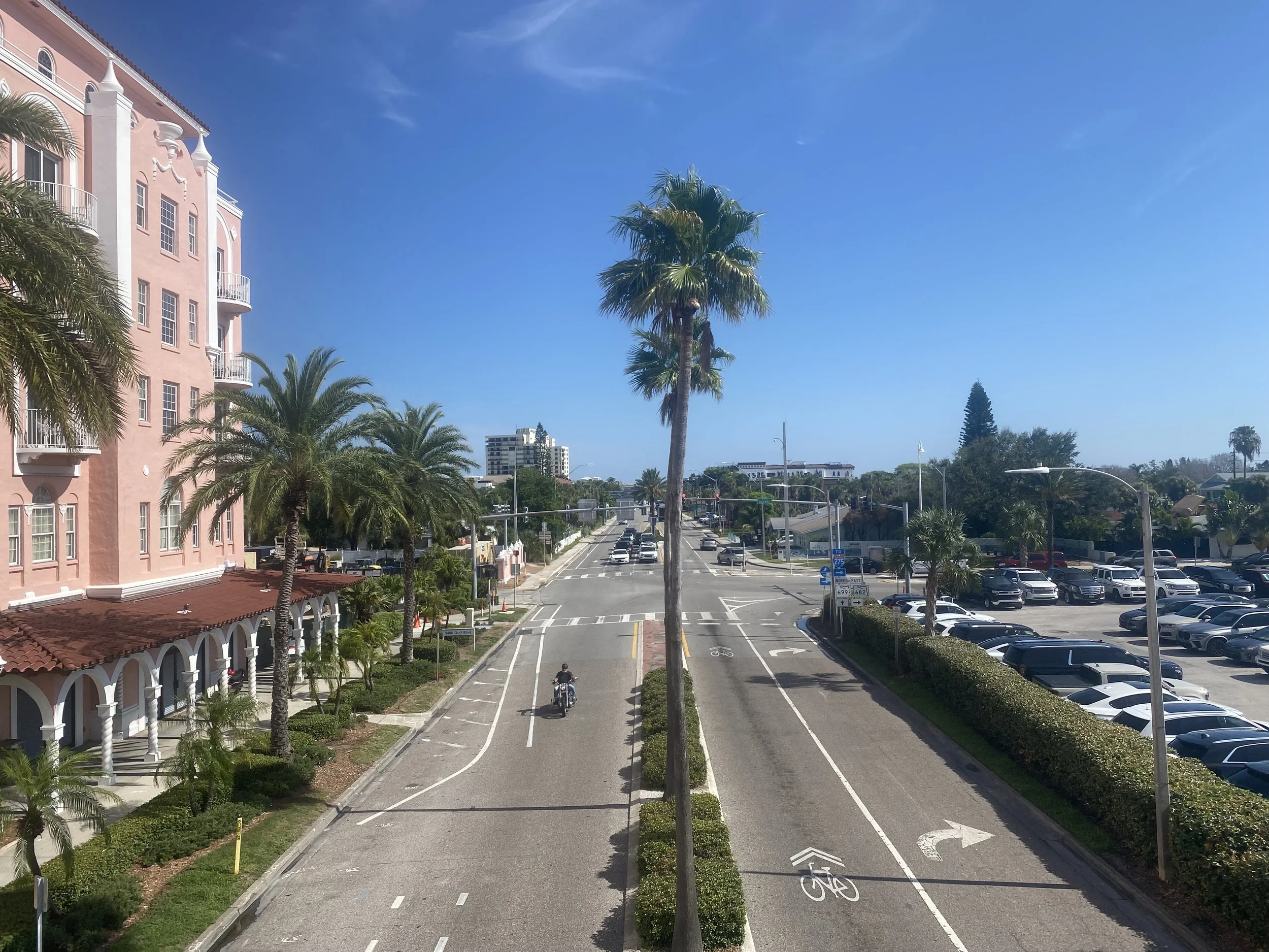 A city street with palm trees, a pink building on the left, cars parked on the right, and a clear blue sky. Gulf boulevard and leading to the prestigious and sought after passe grille waterfront district