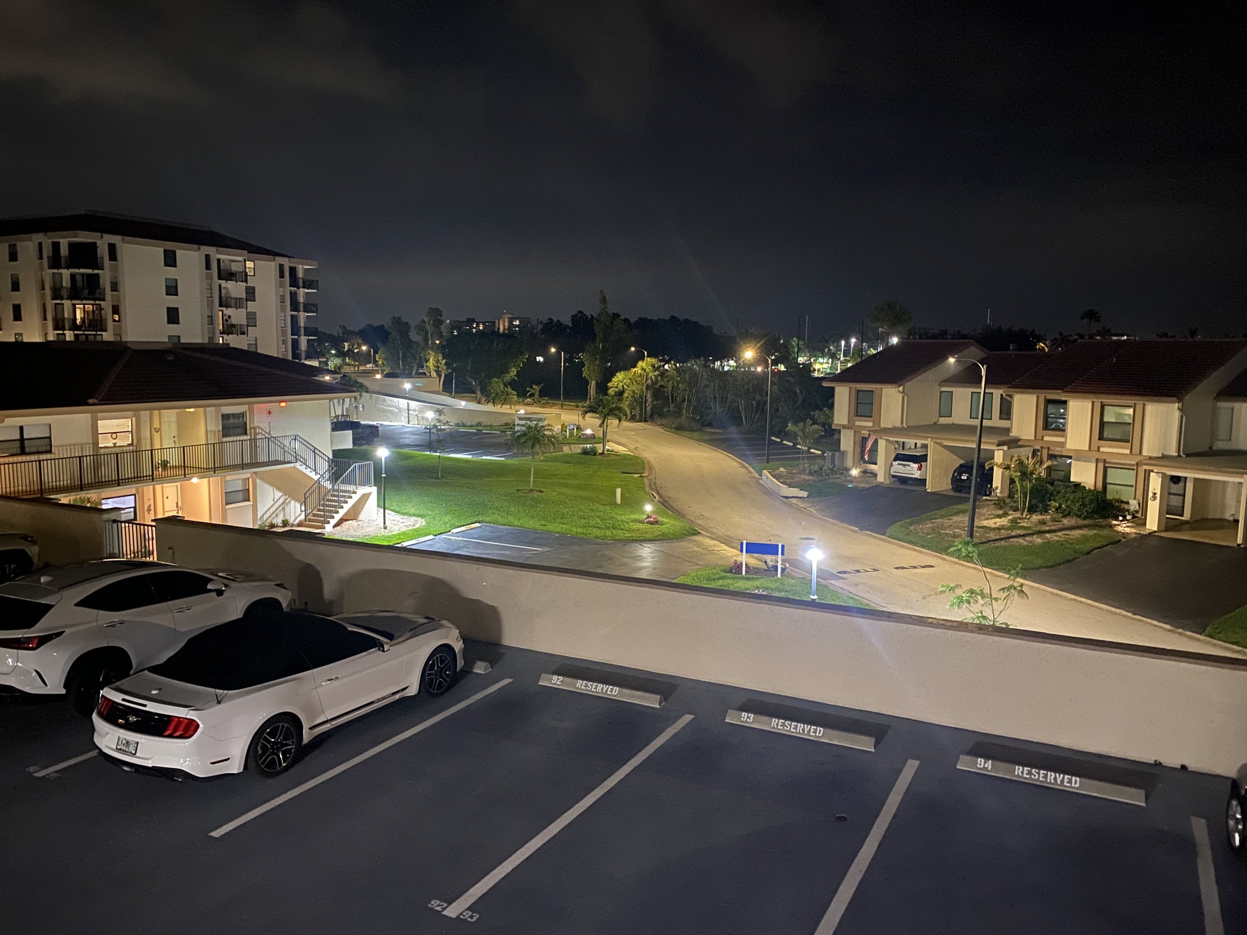 Nighttime view of a residential parking lot with three white cars parked in designated spots labeled 'RESERVED'. Surrounding buildings are apartment complexes with lit windows, and a landscaped area with trees and streetlights illuminates the scene.