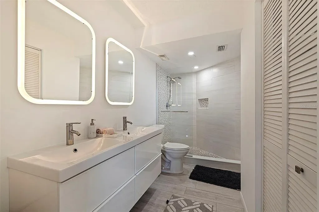Modern white bathroom with double sink vanity, large illuminated mirrors, walk-in shower with bench, and a white toilet.For a luxury experience