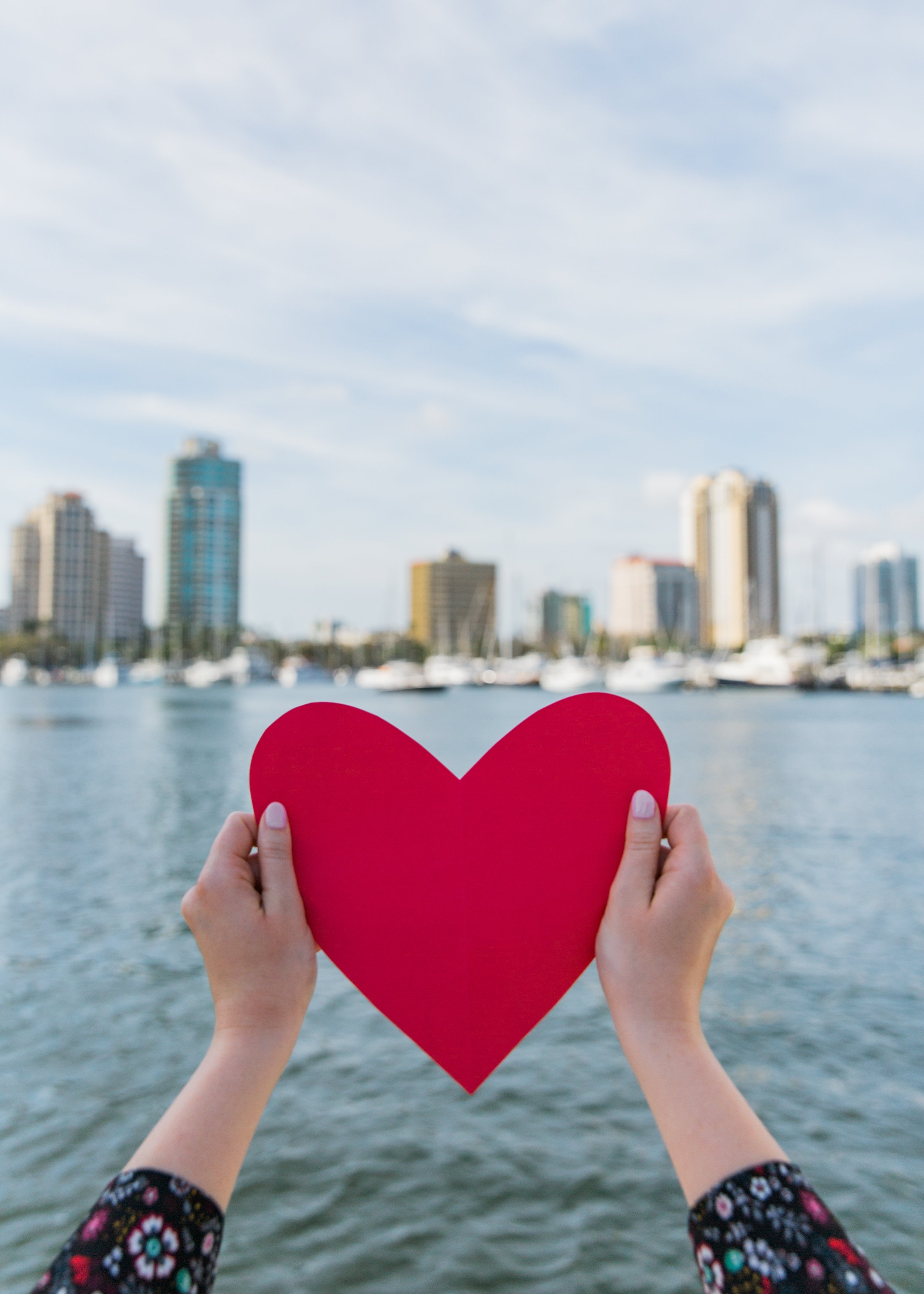 Hands holding a red paper heart in front of a city skyline and water, with a blue sky and clouds in the background representing St Petersburg is somewhere people love to be
