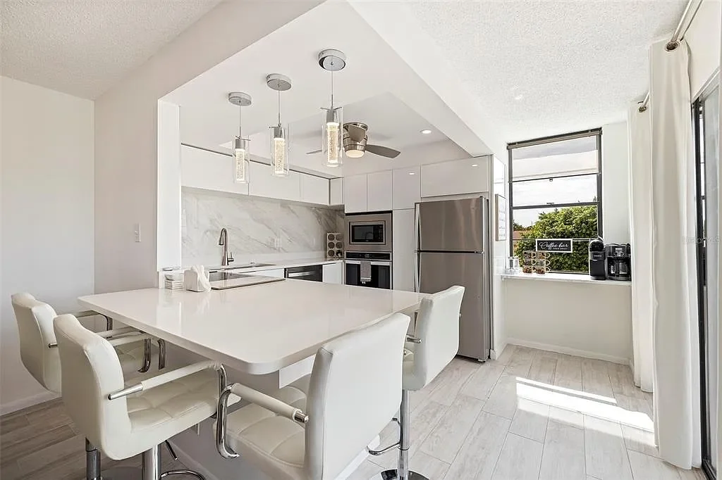 Modern white kitchen with a large island, four white chairs, stainless steel appliances, and a window with curtains.  For a luxury accomodation