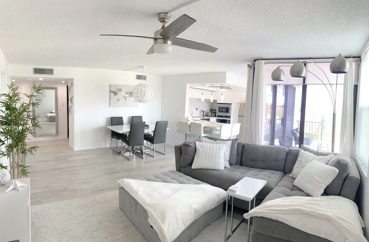 Living room with gray sectional sofa, white pillows, and blanket, next to floor-to-ceiling windows with white curtains, in an open-concept home with dining area and kitchen, light-colored floors, ceiling fan, and balcony.   For a luxury experience