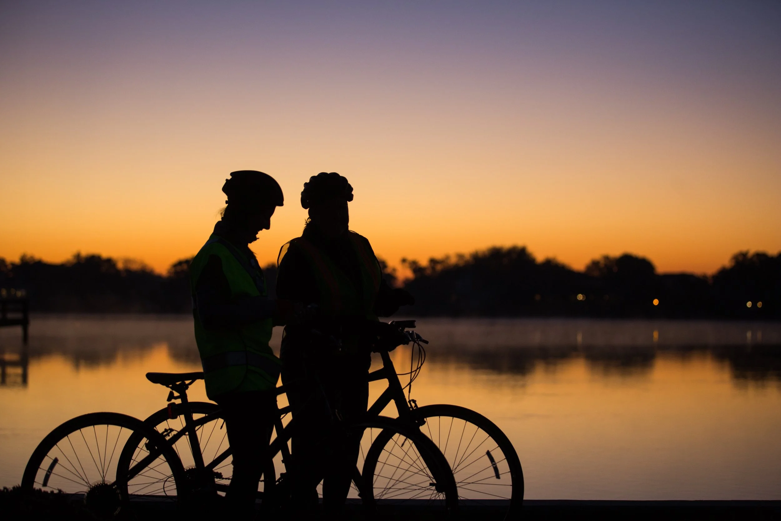 Silhouettes of two people with bicycles, wearing helmets, standing near a body of water at sunset, with trees and a colorful sky in the background.