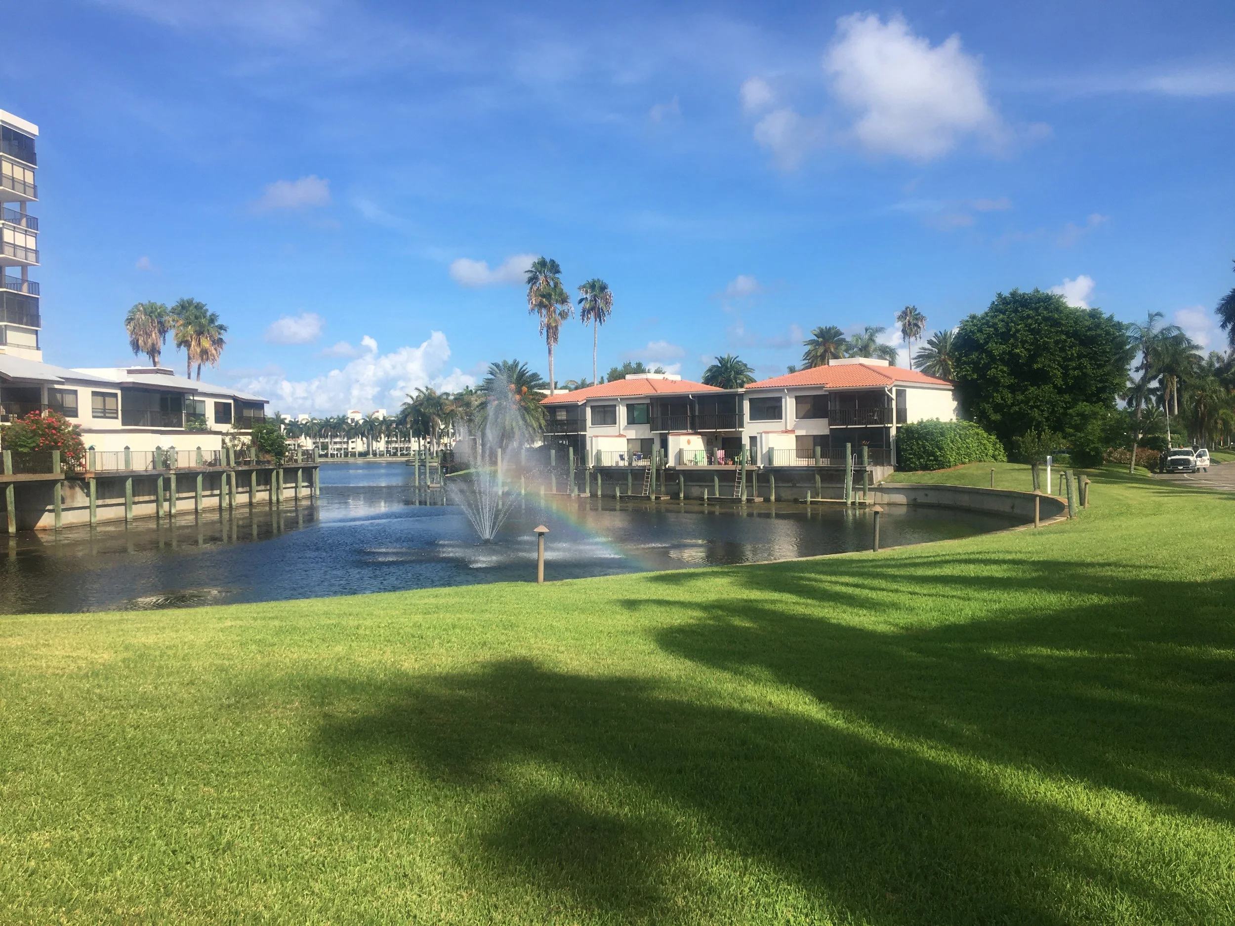 A waterway with a fountain creating a rainbow in the water, surrounded by houses, palm trees, and a grassy foreground under a blue sky with clouds. Beautiful and landscaped grounds and part of a championship golf course