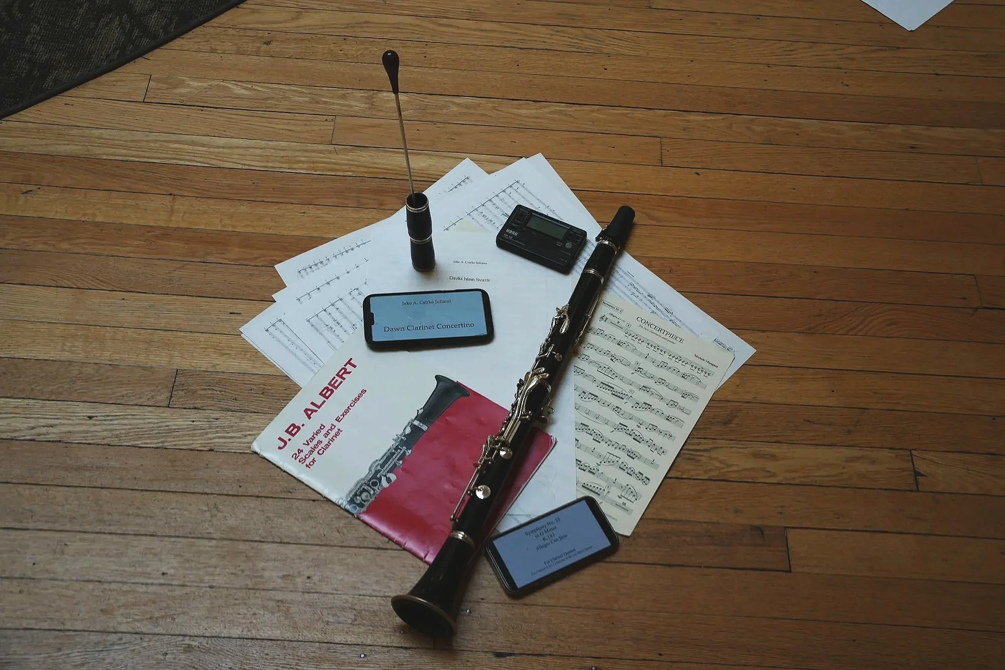 An overhead view of a wooden floor with scattered sheet music, a bass clarinet, a digital tuner, a smartphone displaying music lyrics, and other musical accessories.