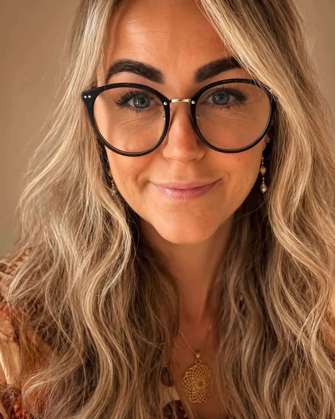 Nærbillede af en kvinde med krøllet, blond hår, store runde briller, smilende, iført øreringe og et halskæde.