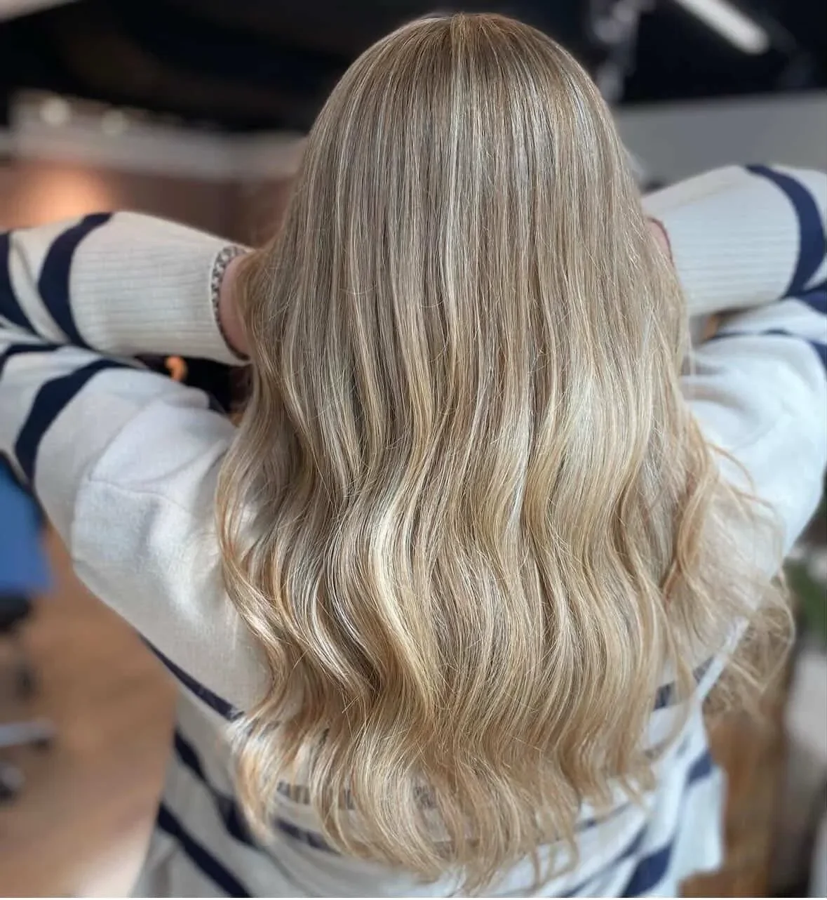 Person med langt, blond, bølget hår set bagfra, med armene hvilede bag hovedet. Babylight farvebehandling hos frisør.
