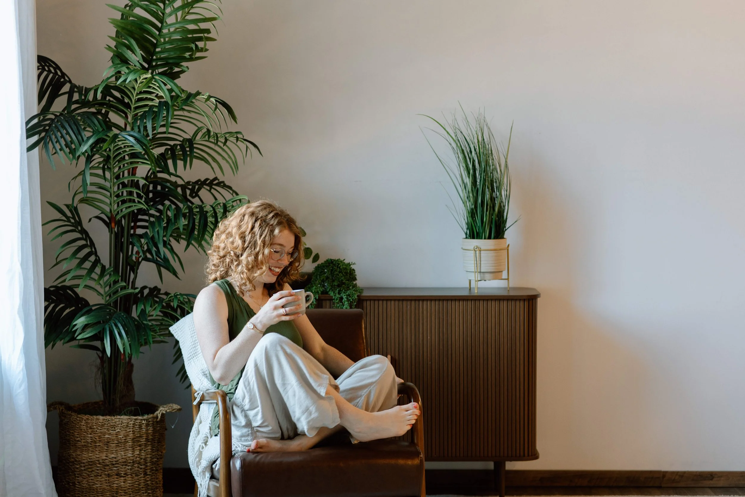 A woman sitting in a brown leather chair indoors, holding a white mug with both hands, smiling and looking down. She has curly blonde hair, wears glasses, a green top, and cream-colored pants. Behind her is a tall green plant with broad leaves, a wicker basket, and a wooden sideboard with a potted plant in a light-colored container. The room has a beige wall and a white curtain on the left side.