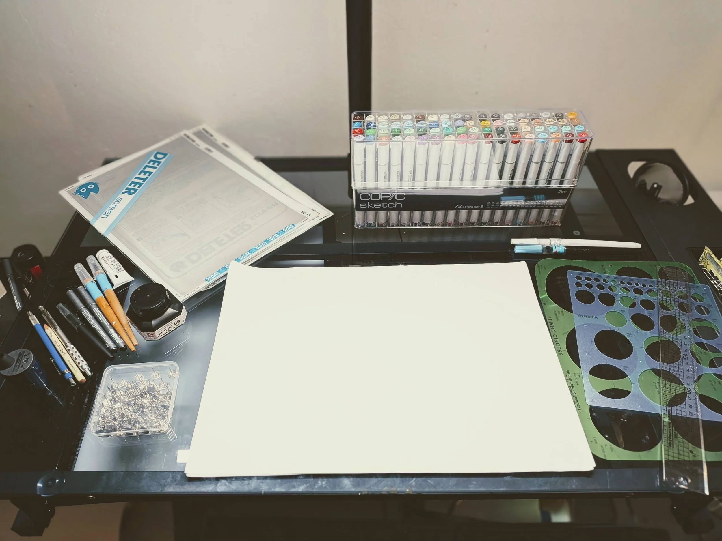 Desk with art supplies including a set of colorful markers, a sheet of paper, various pens and markers, a stencil, a ruler, and a container of small accessories, all arranged on a black table.