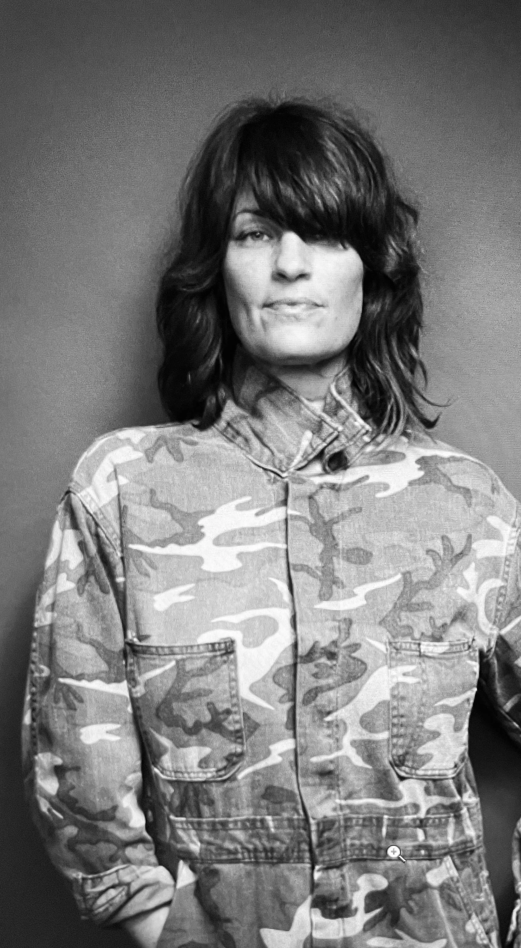 Black and white photo of a woman with shoulder-length wavy hair, wearing a camouflage jacket, standing against a plain wall.