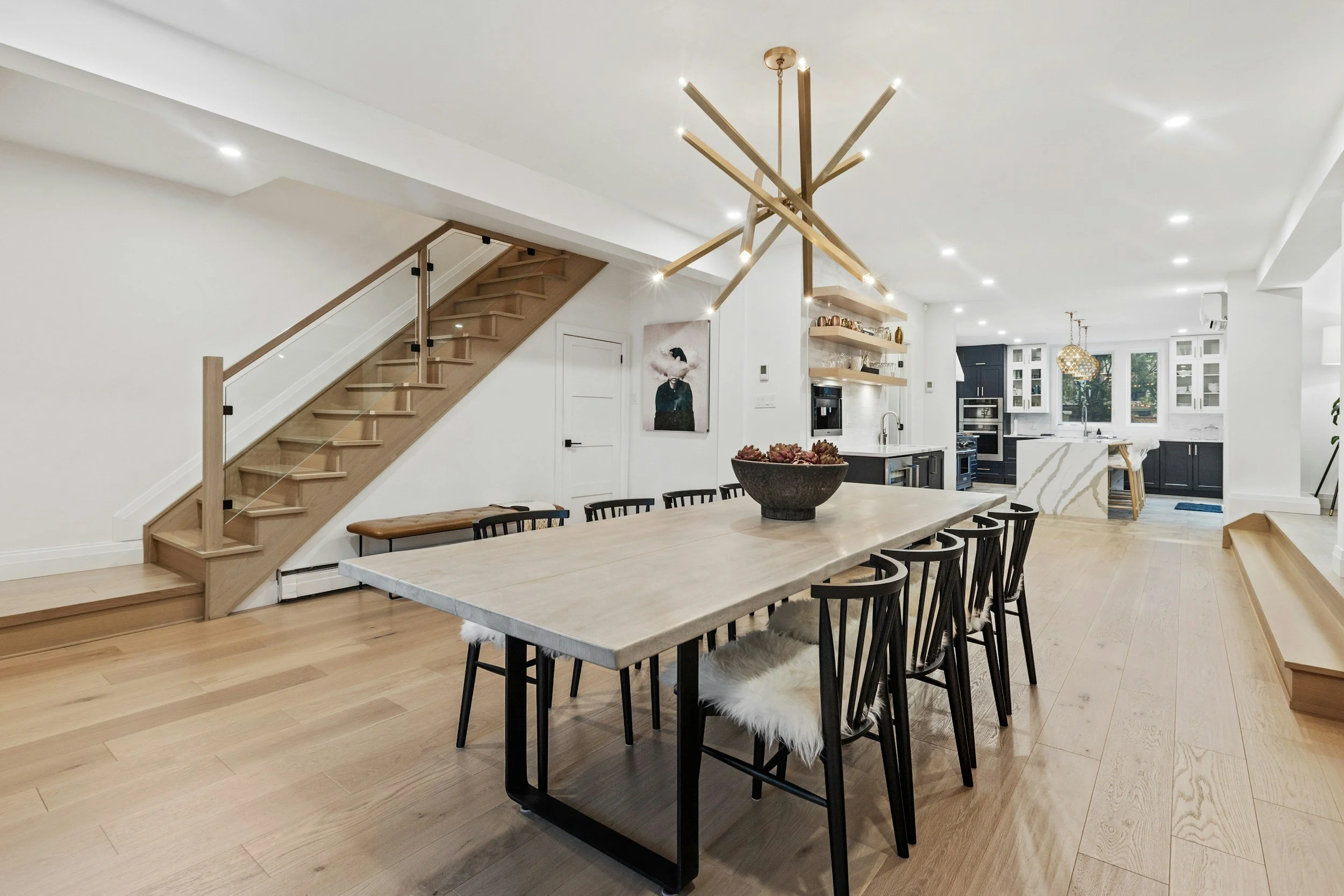 Modern open-concept kitchen and dining area with a large wooden dining table, black chairs with faux fur cushions, a staircase with wooden steps and glass railing, contemporary lighting fixtures, and white walls with built-in shelves.