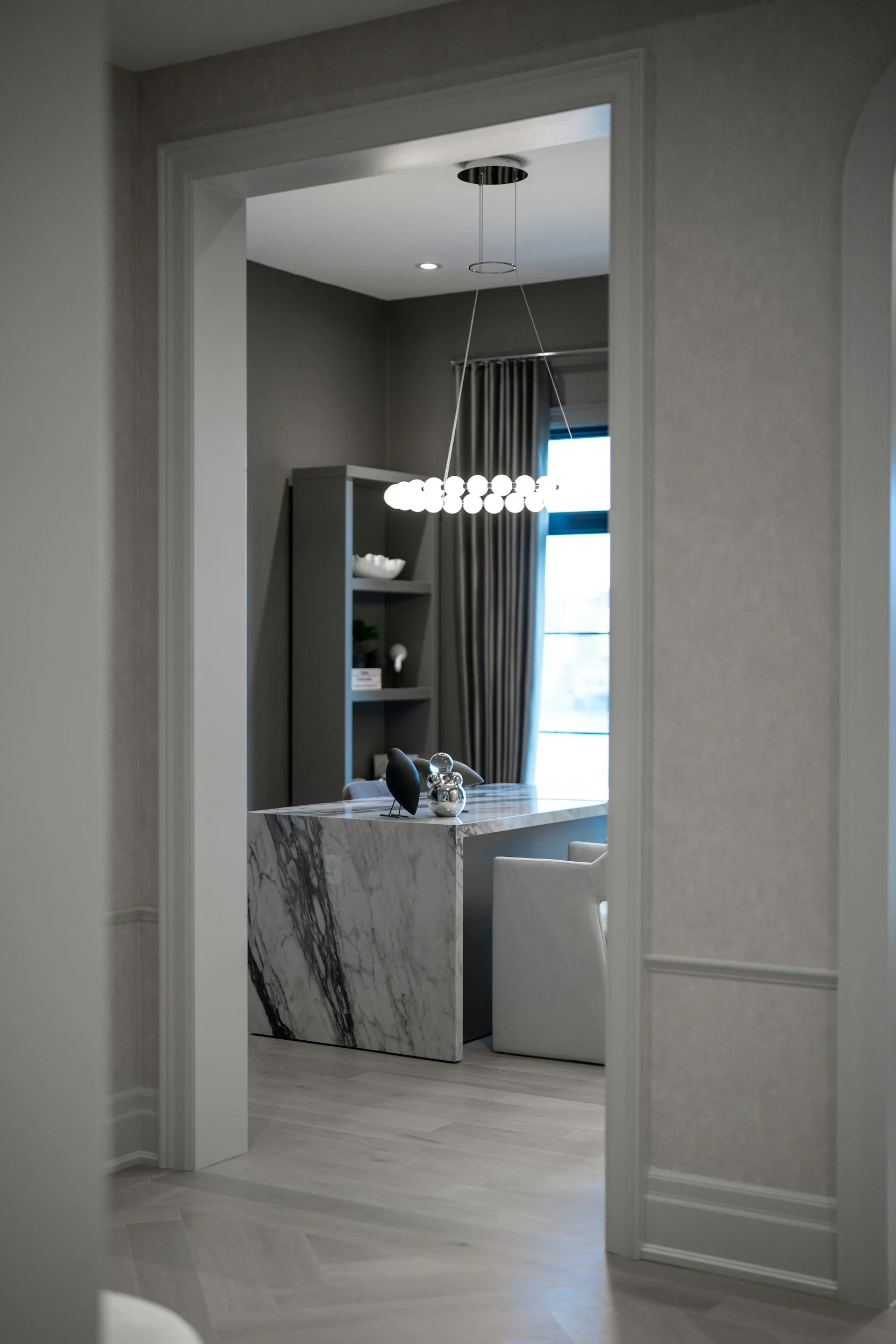 View through a doorway into a dining area with a marble countertop, decorative objects, and a modern hanging light fixture, with a window with curtains in the background.