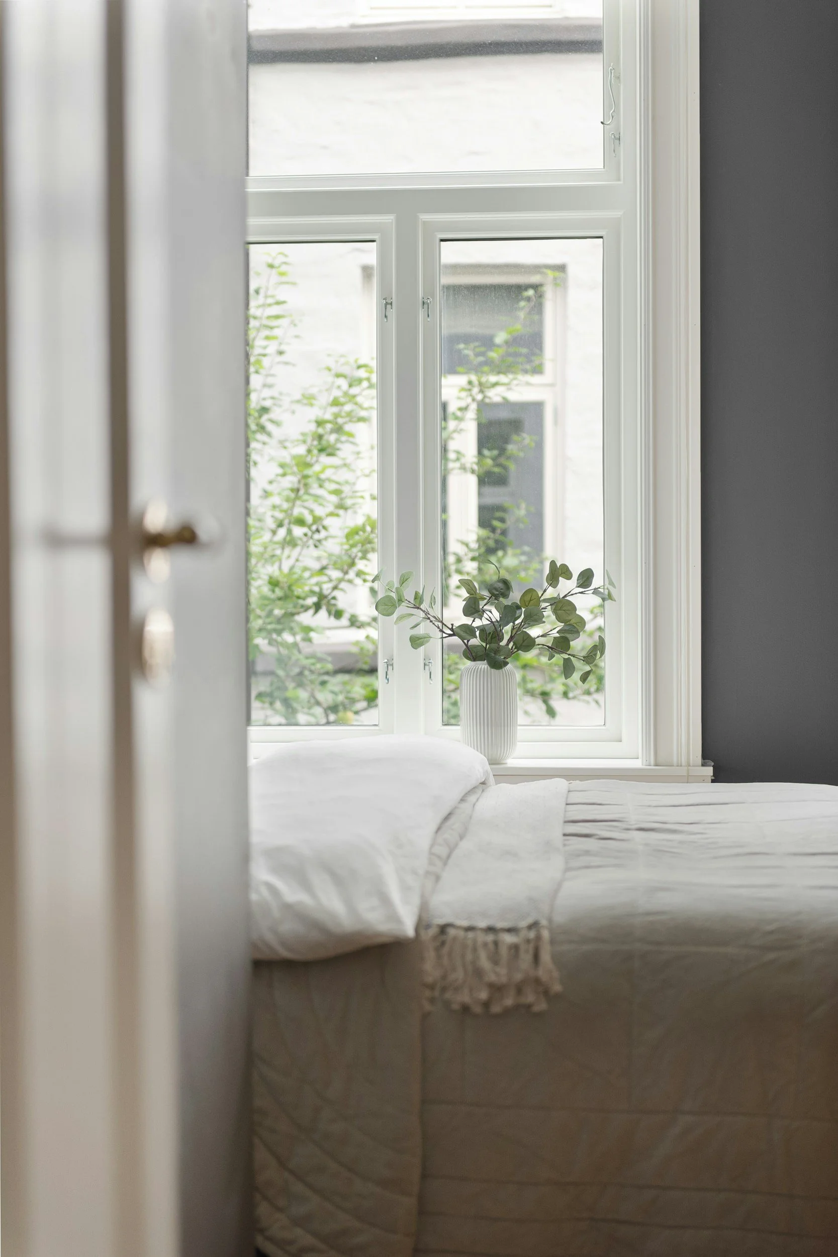 A bedroom seen through an open door showing a bed with white bedding, a window with white frames, a small plant in a white striped vase, and a view of greenery outside.